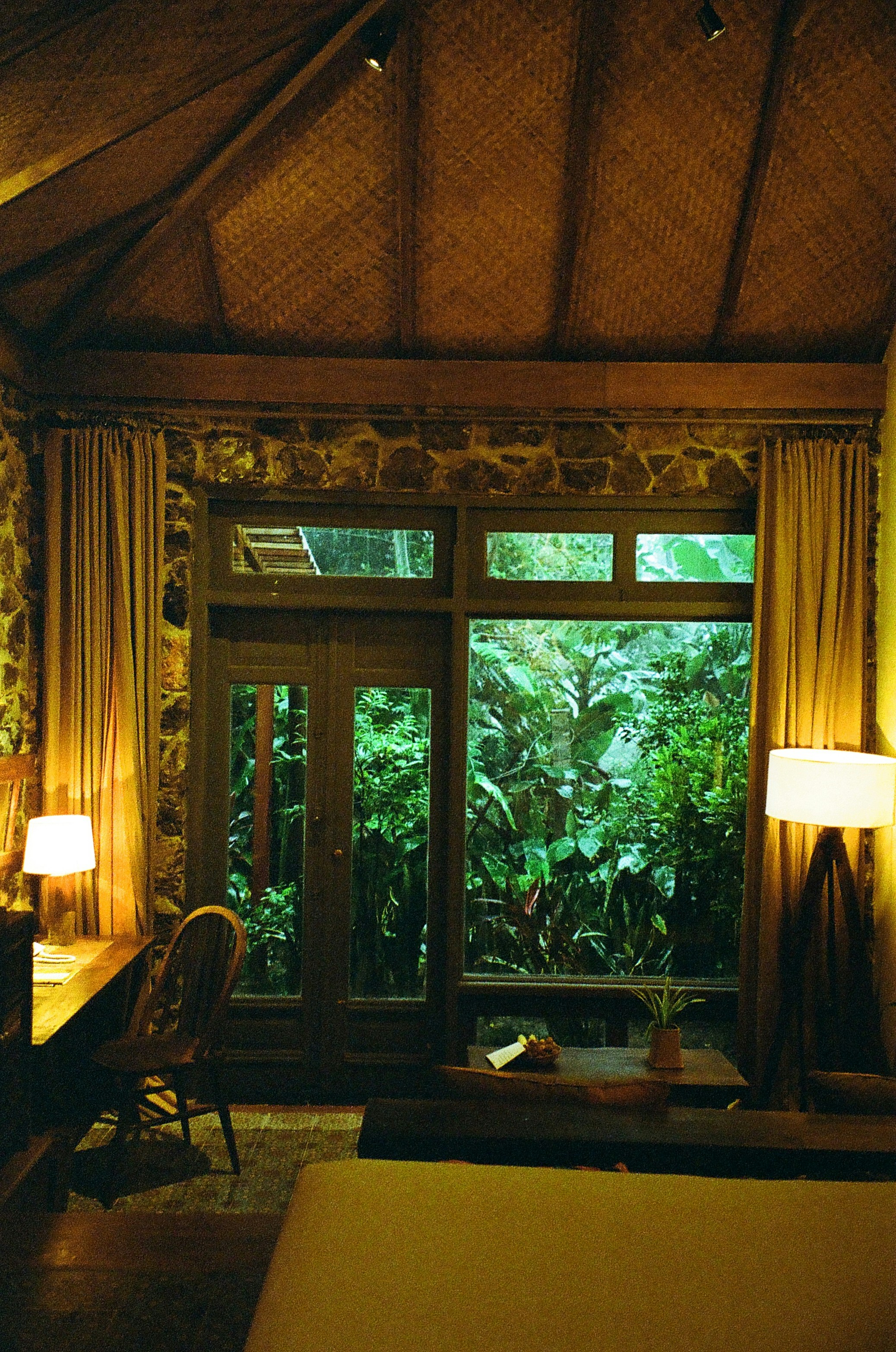 A cozy room with wooden interior features a large window overlooking lush greenery. The room is warmly lit by a table lamp and a floor lamp, creating a relaxing ambiance. A wooden desk with a chair is placed next to the window, and a small table holds a plant beside the windowsill. The room has a thatched ceiling and stone walls.