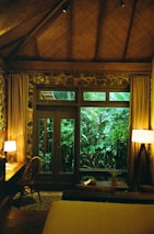 A cozy room with wooden interior features a large window overlooking lush greenery. The room is warmly lit by a table lamp and a floor lamp, creating a relaxing ambiance. A wooden desk with a chair is placed next to the window, and a small table holds a plant beside the windowsill. The room has a thatched ceiling and stone walls.