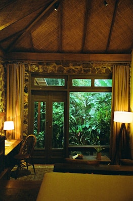 A cozy room with wooden interior features a large window overlooking lush greenery. The room is warmly lit by a table lamp and a floor lamp, creating a relaxing ambiance. A wooden desk with a chair is placed next to the window, and a small table holds a plant beside the windowsill. The room has a thatched ceiling and stone walls.