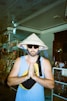 A person stands with their hands together in a gesture of prayer or greeting. They are wearing a traditional Vietnamese conical hat with the word 'Vietnam' written on it and sunglasses. The person is also wearing a blue sleeveless shirt and a crossbody bag. The background features various items on shelves, possibly in a shop or market.