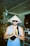 A person stands with their hands together in a gesture of prayer or greeting. They are wearing a traditional Vietnamese conical hat with the word 'Vietnam' written on it and sunglasses. The person is also wearing a blue sleeveless shirt and a crossbody bag. The background features various items on shelves, possibly in a shop or market.