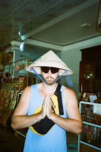 A person stands with their hands together in a gesture of prayer or greeting. They are wearing a traditional Vietnamese conical hat with the word 'Vietnam' written on it and sunglasses. The person is also wearing a blue sleeveless shirt and a crossbody bag. The background features various items on shelves, possibly in a shop or market.