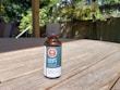 A small amber glass bottle with a black cap labeled 'Lofi THC CBD Oil 1500' sits on a wooden table. The label features a red hexagon with a white cannabis leaf symbol and the words 'THC' and 'CBD oil / 1500'. Sunlight creates shadows on the table, and there are green leafy plants and a fence in the soft-focus background.