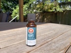 A small amber glass bottle with a black cap labeled 'Lofi THC CBD Oil 1500' sits on a wooden table. The label features a red hexagon with a white cannabis leaf symbol and the words 'THC' and 'CBD oil / 1500'. Sunlight creates shadows on the table, and there are green leafy plants and a fence in the soft-focus background.