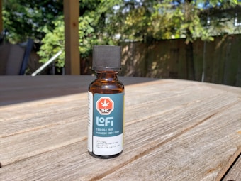 A small amber glass bottle with a black cap labeled 'Lofi THC CBD Oil 1500' sits on a wooden table. The label features a red hexagon with a white cannabis leaf symbol and the words 'THC' and 'CBD oil / 1500'. Sunlight creates shadows on the table, and there are green leafy plants and a fence in the soft-focus background.