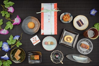 An assortment of traditional Japanese sweets artistically arranged on a dark, textured surface. Each sweet is placed in a unique dish, ranging from round and square plates to leaf-shaped trays. Vibrant artificial morning glory flowers with green leaves accentuate the setup. In the center, a wrapped gift box tied with a ribbon suggests a special occasion. Nearby, a card with Japanese text and a logo complements the arrangement. The scene combines delicacies like a roll cake, colorful dango, and various pastries, reflecting a diverse culinary display.