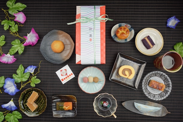 An assortment of traditional Japanese sweets artistically arranged on a dark, textured surface. Each sweet is placed in a unique dish, ranging from round and square plates to leaf-shaped trays. Vibrant artificial morning glory flowers with green leaves accentuate the setup. In the center, a wrapped gift box tied with a ribbon suggests a special occasion. Nearby, a card with Japanese text and a logo complements the arrangement. The scene combines delicacies like a roll cake, colorful dango, and various pastries, reflecting a diverse culinary display.