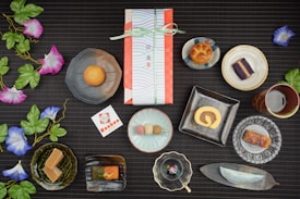 An assortment of traditional Japanese sweets artistically arranged on a dark, textured surface. Each sweet is placed in a unique dish, ranging from round and square plates to leaf-shaped trays. Vibrant artificial morning glory flowers with green leaves accentuate the setup. In the center, a wrapped gift box tied with a ribbon suggests a special occasion. Nearby, a card with Japanese text and a logo complements the arrangement. The scene combines delicacies like a roll cake, colorful dango, and various pastries, reflecting a diverse culinary display.