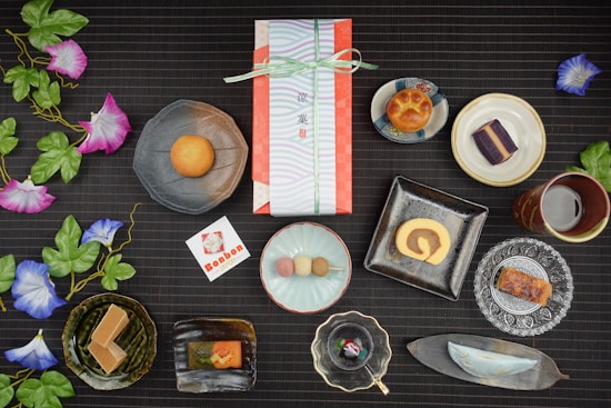 An assortment of traditional Japanese sweets artistically arranged on a dark, textured surface. Each sweet is placed in a unique dish, ranging from round and square plates to leaf-shaped trays. Vibrant artificial morning glory flowers with green leaves accentuate the setup. In the center, a wrapped gift box tied with a ribbon suggests a special occasion. Nearby, a card with Japanese text and a logo complements the arrangement. The scene combines delicacies like a roll cake, colorful dango, and various pastries, reflecting a diverse culinary display.