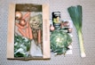 A collection of fresh vegetables including onions, carrots, celeriac, leeks, a leafy green vegetable, and a can of organic coconut milk are arranged on a patterned surface. The items are organized with some inside a cardboard box labeled 'Feel Good Farming' and others placed alongside a magazine feature.