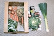 A collection of fresh vegetables including onions, carrots, celeriac, leeks, a leafy green vegetable, and a can of organic coconut milk are arranged on a patterned surface. The items are organized with some inside a cardboard box labeled 'Feel Good Farming' and others placed alongside a magazine feature.