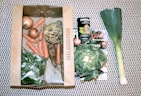 A collection of fresh vegetables including onions, carrots, celeriac, leeks, a leafy green vegetable, and a can of organic coconut milk are arranged on a patterned surface. The items are organized with some inside a cardboard box labeled 'Feel Good Farming' and others placed alongside a magazine feature.