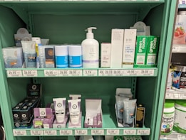 A store shelf filled with various health and wellness products, including lotions, creams, and lip balms. The shelf features brands like Charlotte's Web, Social, Joy, and Green Roads. Products are labeled with prices and organized neatly by brand. The shelf is painted in a light green color, complementing the packaging design of many products.
