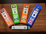 Four CBD vape pen boxes are arranged on a wooden surface. Each box is brightly colored, featuring different flavors: Strawberry Lemonade, Fresh Mint, Tropic Breeze, and Blue Raspberry. The boxes indicate 500mg of CBD per pen. A single white vape pen with the CBD logo lies horizontally below the boxes.