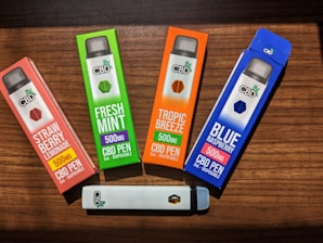 Four CBD vape pen boxes are arranged on a wooden surface. Each box is brightly colored, featuring different flavors: Strawberry Lemonade, Fresh Mint, Tropic Breeze, and Blue Raspberry. The boxes indicate 500mg of CBD per pen. A single white vape pen with the CBD logo lies horizontally below the boxes.