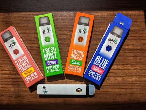 Four CBD vape pen boxes are arranged on a wooden surface. Each box is brightly colored, featuring different flavors: Strawberry Lemonade, Fresh Mint, Tropic Breeze, and Blue Raspberry. The boxes indicate 500mg of CBD per pen. A single white vape pen with the CBD logo lies horizontally below the boxes.