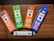 Four CBD vape pen boxes are arranged on a wooden surface. Each box is brightly colored, featuring different flavors: Strawberry Lemonade, Fresh Mint, Tropic Breeze, and Blue Raspberry. The boxes indicate 500mg of CBD per pen. A single white vape pen with the CBD logo lies horizontally below the boxes.