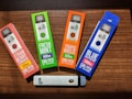 Four CBD vape pen boxes are arranged on a wooden surface. Each box is brightly colored, featuring different flavors: Strawberry Lemonade, Fresh Mint, Tropic Breeze, and Blue Raspberry. The boxes indicate 500mg of CBD per pen. A single white vape pen with the CBD logo lies horizontally below the boxes.