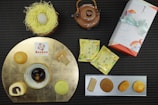 A selection of Japanese desserts is elegantly arranged on a dark, textured background. There are various sweets on a gold circular plate, including a spiral pastry and a black confection. A brown teapot with an intricate handle is placed next to a wrapped rectangular gift adorned with a light blue ribbon featuring fish illustrations. Two yellow tea packets with floral designs and a decorative nest containing a round white dessert complete the display.
