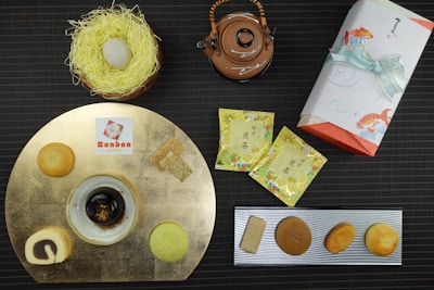 A selection of Japanese desserts is elegantly arranged on a dark, textured background. There are various sweets on a gold circular plate, including a spiral pastry and a black confection. A brown teapot with an intricate handle is placed next to a wrapped rectangular gift adorned with a light blue ribbon featuring fish illustrations. Two yellow tea packets with floral designs and a decorative nest containing a round white dessert complete the display.