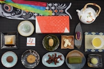 A variety of Japanese sweets and desserts are arranged neatly on a black mat. There are colorful elements, such as a koi-shaped decoration at the top, a red-patterned box in the center, and a white teapot with floral designs at the top right. Plates and bowls hold different confections, including mochi, jellies, and a small cup of green tea.