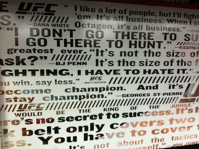 A wall filled with motivational quotes associated with the UFC. The text includes statements from notable individuals like Dana White, BJ Penn, and Georges St-Pierre, emphasizing themes of competition, strength, and success. The typography is bold and varied in size, with a mixture of black, gray, and some red on a white background.