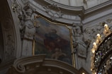 An intricately decorated interior features a classical painting surrounded by ornate sculptural elements and gilded accents. The painting depicts a seated figure in historical attire with a cherub nearby. On either side, detailed sculptures of robed figures are positioned against a gracefully arched architectural backdrop, adorned with elaborate patterns.