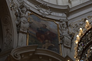 An intricately decorated interior features a classical painting surrounded by ornate sculptural elements and gilded accents. The painting depicts a seated figure in historical attire with a cherub nearby. On either side, detailed sculptures of robed figures are positioned against a gracefully arched architectural backdrop, adorned with elaborate patterns.