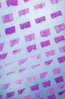 Brightly colored papel picado, traditional Mexican cut-out paper banners, are strung across the sky. The banners feature intricate designs and some bear the text 'CENTRO HISTORICO,' with a backdrop of a pale blue sky scattered with a few clouds.