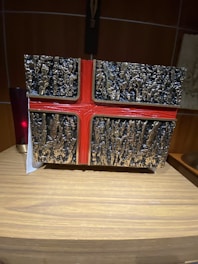 A textured object with a decorative cross design sits on a wooden surface. The object has a rough, black and silver surface, contrasted by a smooth red cross overlay. A dark red cylindrical object can be seen partially in the background.