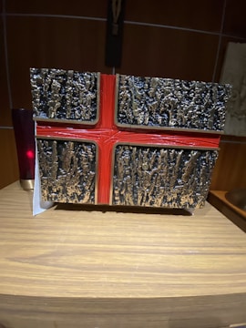 A textured object with a decorative cross design sits on a wooden surface. The object has a rough, black and silver surface, contrasted by a smooth red cross overlay. A dark red cylindrical object can be seen partially in the background.