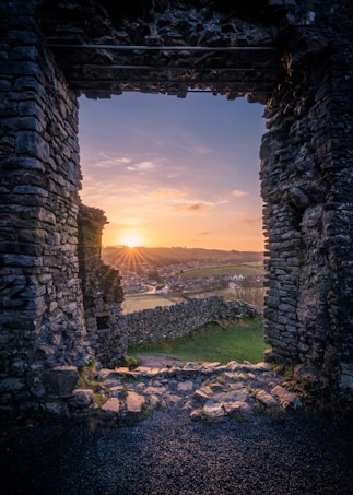 A sunset view is framed by the stone archway of an ancient ruin. The sun casts a warm glow over a serene landscape, including rolling hills, a stone wall, and buildings in the distance. The sky features a gradient of colors from oranges and yellows near the horizon to soft blues higher up.