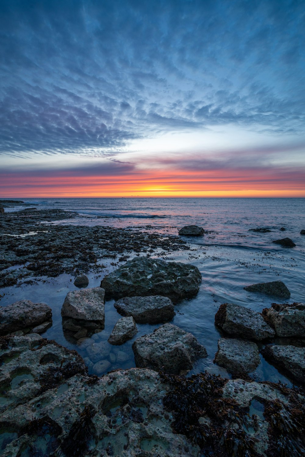 the sun is setting over the ocean with rocks in the foreground