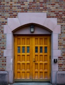 A wooden double door with a natural finish set within an arched stone doorway. The door has multiple panels and small glass windows, surrounded by a wall of red and beige bricks. An access control keypad is mounted on the right, above which is a rectangular light fixture.
