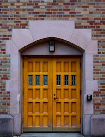 A wooden double door with a natural finish set within an arched stone doorway. The door has multiple panels and small glass windows, surrounded by a wall of red and beige bricks. An access control keypad is mounted on the right, above which is a rectangular light fixture.