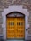 A wooden double door with a natural finish set within an arched stone doorway. The door has multiple panels and small glass windows, surrounded by a wall of red and beige bricks. An access control keypad is mounted on the right, above which is a rectangular light fixture.