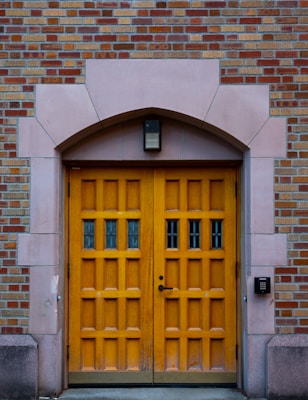 A wooden double door with a natural finish set within an arched stone doorway. The door has multiple panels and small glass windows, surrounded by a wall of red and beige bricks. An access control keypad is mounted on the right, above which is a rectangular light fixture.