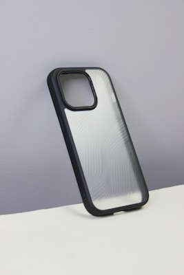 A smartphone case with a sleek, semi-transparent back featuring curvy line patterns. The case has a black border and is displayed against a plain, grayish wall with a white surface at the bottom.