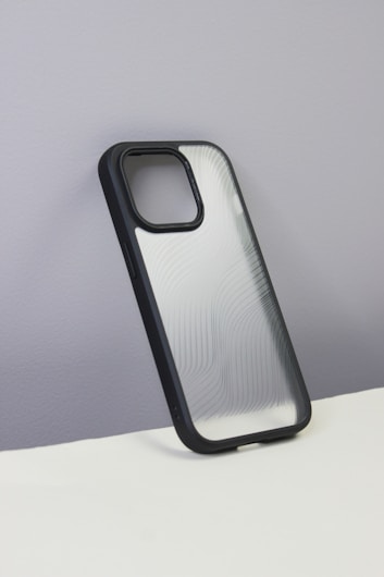 A smartphone case with a sleek, semi-transparent back featuring curvy line patterns. The case has a black border and is displayed against a plain, grayish wall with a white surface at the bottom.