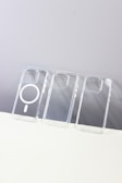 Three transparent phone cases are lined up vertically on a flat surface. The middle case has a MagSafe ring, while the others are plain. The background is split between a light gray and white surface.
