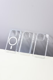 Three transparent phone cases are lined up vertically on a flat surface. The middle case has a MagSafe ring, while the others are plain. The background is split between a light gray and white surface.