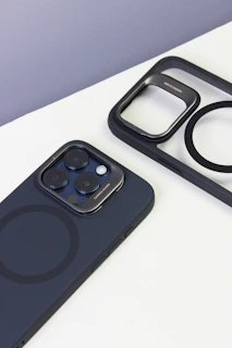A smartphone with a black case and visible camera lenses is placed on a white surface. Nearby, there is a matching black phone case with a cutout pattern, showing the phone's MagSafe circle design.