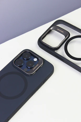 A smartphone with a black case and visible camera lenses is placed on a white surface. Nearby, there is a matching black phone case with a cutout pattern, showing the phone's MagSafe circle design.
