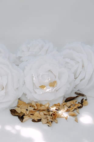 Large white paper flowers are arranged near a cluster of dry, brown leaves. The setting has soft lighting with gentle shadows, giving a serene and calm atmosphere.