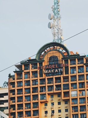 A high-rise building with multiple balconies and a large sign that reads Citadel Inn Makati. The structure has an earthy orange and brown color with a prominent tower featuring several satellite dishes on top.