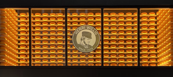 A display of numerous rounds of cheese neatly stacked on shelves is visible. The wall is backlit with warm yellow lighting, creating a harmonious and inviting atmosphere. In the center, there is a logo with the text 'Henri Willig Since 1974', featuring a profile image.
