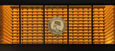 A display of numerous rounds of cheese neatly stacked on shelves is visible. The wall is backlit with warm yellow lighting, creating a harmonious and inviting atmosphere. In the center, there is a logo with the text 'Henri Willig Since 1974', featuring a profile image.