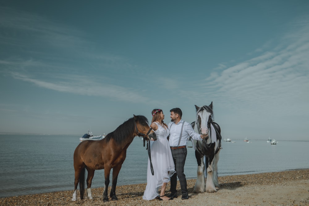 a couple standing next to two horses on a beach
