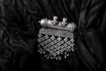 An intricate silver pendant lies on a textured black fabric background. The pendant features detailed patterns with floral and geometric motifs, adorned with small dangling beads.