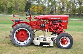 A red vintage tractor is parked on a grassy field. The tractor has large rear wheels and smaller front wheels. It features detailed mechanical components and metal parts, reflecting its classic design. The background shows a line of trees with green and yellow foliage, suggesting a rural setting.